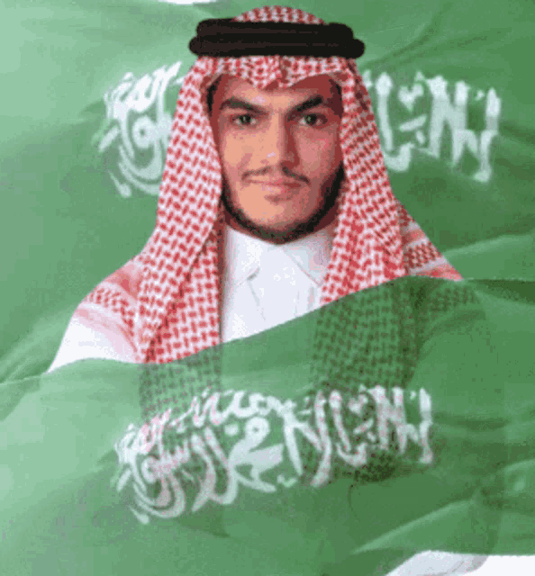 a man in a keffiyeh is behind a green flag that says ' saudi arabia ' on it