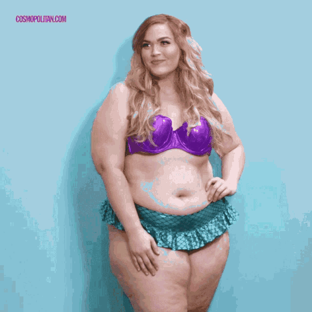 a woman in a purple bikini is standing in front of a blue wall with cosmopolitan.com written on the bottom