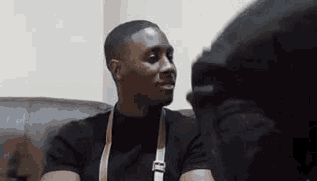 a man in a black shirt is sitting on a couch talking to someone .