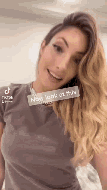 a woman wearing a grey t-shirt and a necklace says " now look at this " on a tiktok video