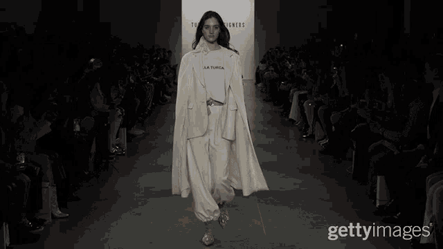 a model walks down a runway wearing a white coat and a white t-shirt that says la turrica