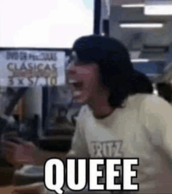 a man is sitting at a table with his mouth open and the word queee is on his shirt .