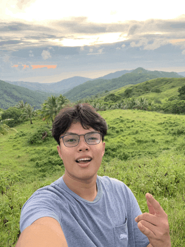 a young man wearing glasses takes a selfie in front of a grassy hillside