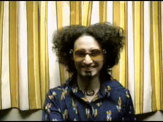 a man with curly hair and sunglasses is smiling in front of a striped curtain