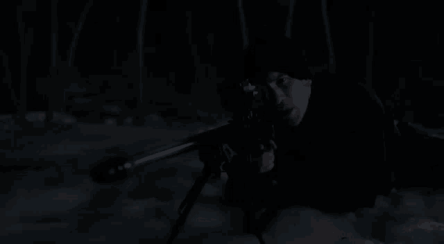 a man is laying in the snow holding a sniper rifle in a dark forest .