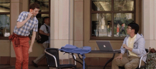 a man talking on a cell phone while another man sits at a table with a laptop