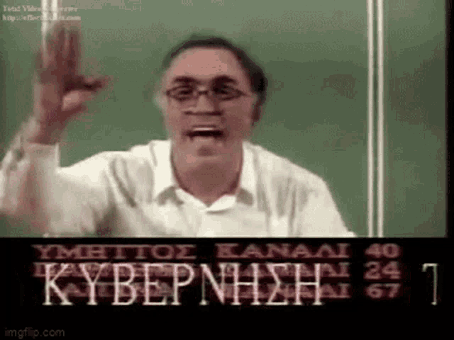 a man wearing glasses is giving a speech in front of a green board with greek writing on it .