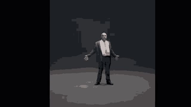 a man in a suit and tie stands in a dark room with his arms outstretched