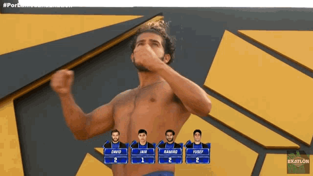a shirtless man stands in front of a wall with a scoreboard for exatlon