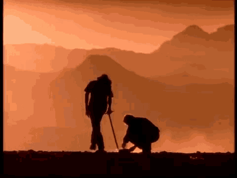a man with a cane is walking next to another man with a shovel in front of a mountain .