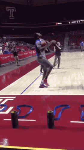 a basketball player jumps in the air on a court with a t on the wall behind him