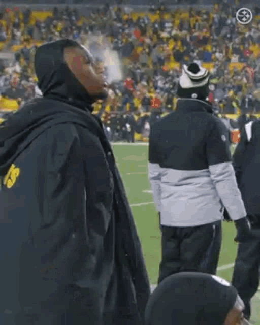 a man is smoking a cigarette on a football field while wearing a hooded jacket .