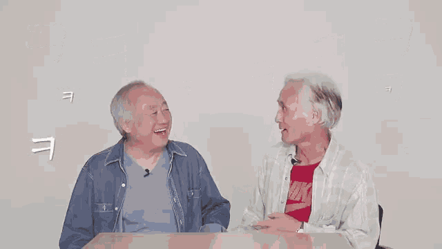 two older men are sitting at a table talking to each other and laughing .