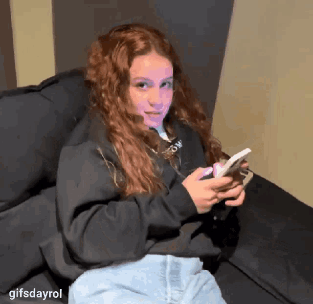 a girl with red hair is sitting on a couch looking at her phone with gifsdayrol written below her