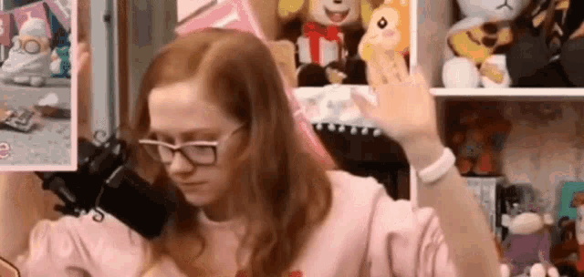 a girl wearing glasses is sitting in front of a microphone in a room filled with stuffed animals .