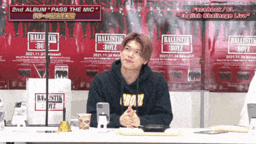 a man in a ballistik boyz hoodie sits at a table in front of posters