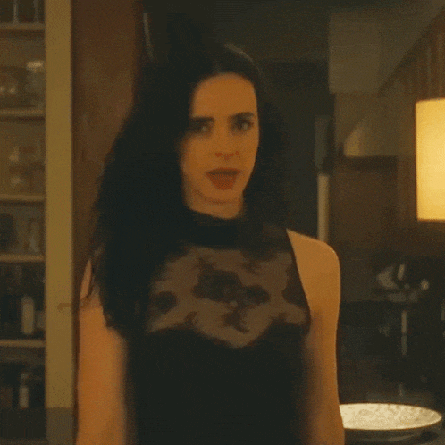 a woman in a black lace top is standing in a kitchen looking at the camera .