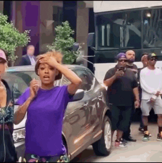 a woman in a purple shirt is standing in front of a group of people standing around a car .