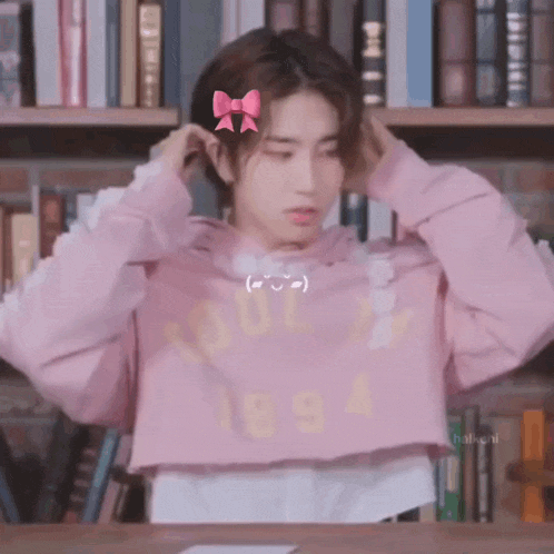 a young boy wearing a pink crop top and a pink bow in his hair is sitting in front of a bookshelf .