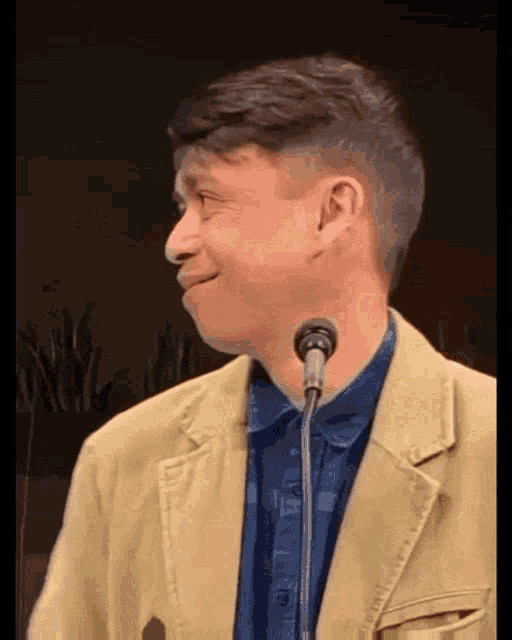 a man wearing a tan jacket and a blue shirt stands in front of a microphone