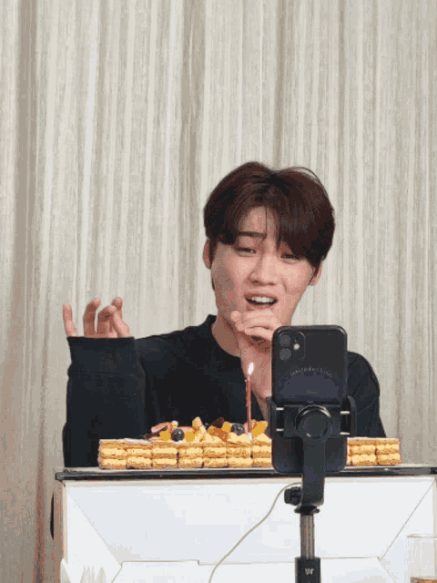 a young man looks at his phone while holding a candle in front of a birthday cake
