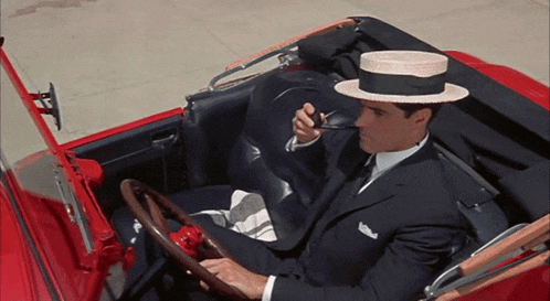 a man in a black suit and hat is driving a red car