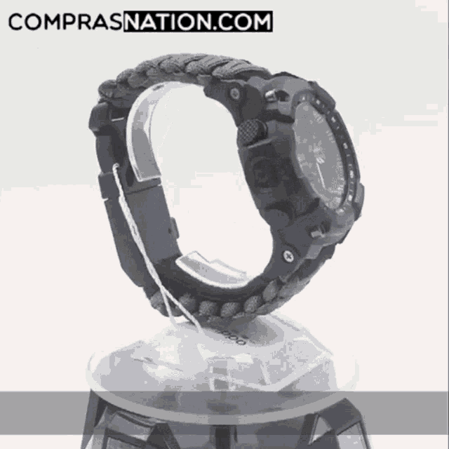 a watch with a paracord band sits on a display stand with the website comprasnation.com written on the bottom