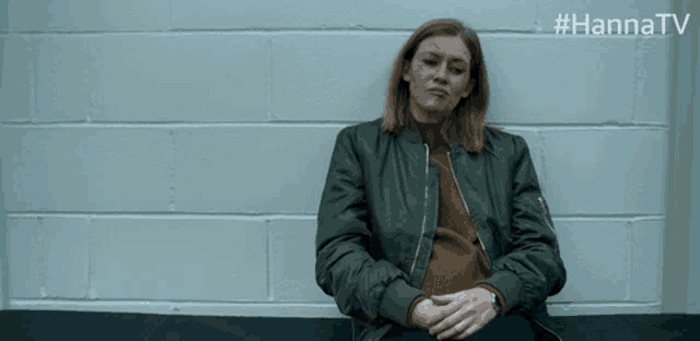 a woman in a green jacket sits in front of a brick wall