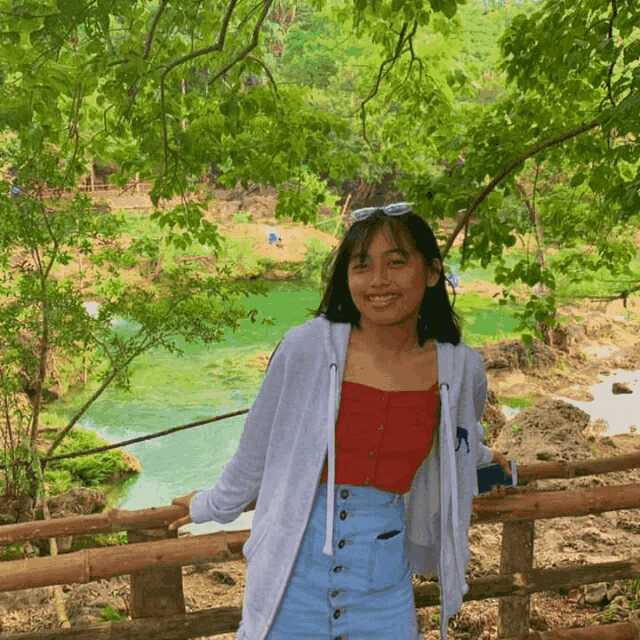 a girl wearing a red top and a white jacket stands in front of a river