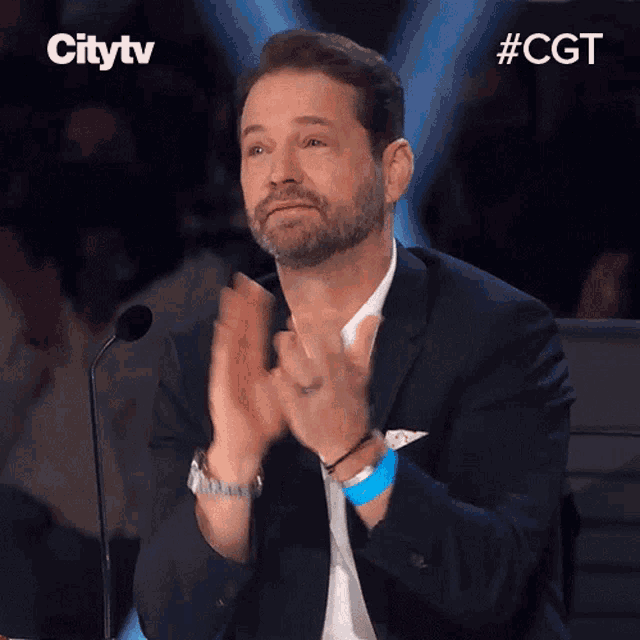 a man in a suit applauds in front of a microphone with the hashtag #cgt on the bottom