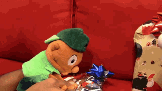 a stuffed animal wearing a green hat is opening a christmas present on a red couch