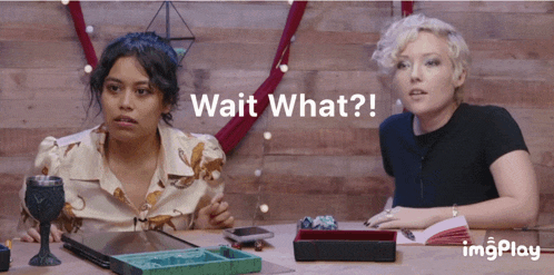 two women are sitting at a table with the words wait what on the bottom