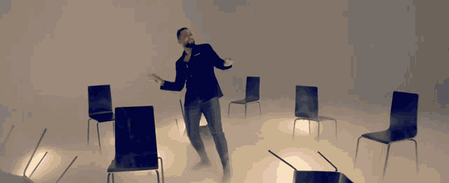 a man in a suit stands in a room with chairs flying around him