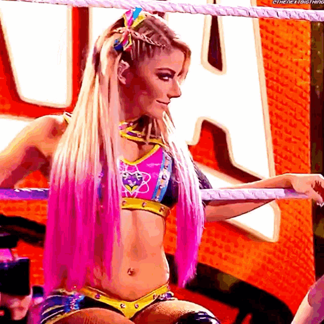a woman with pink hair is sitting in a wrestling ring with a large letter h in the background