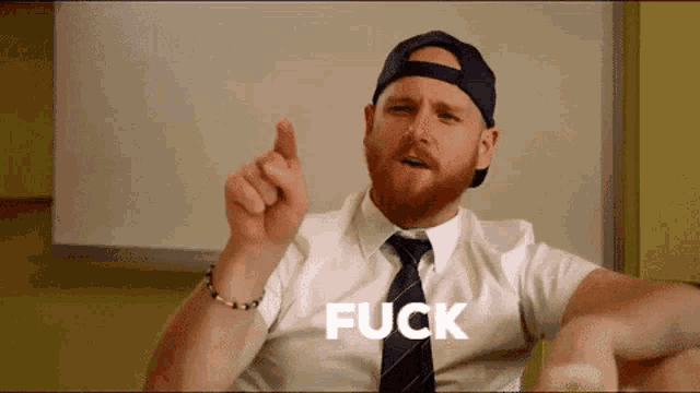 a man with a beard wearing a hat and tie is pointing up with the word fuck behind him .