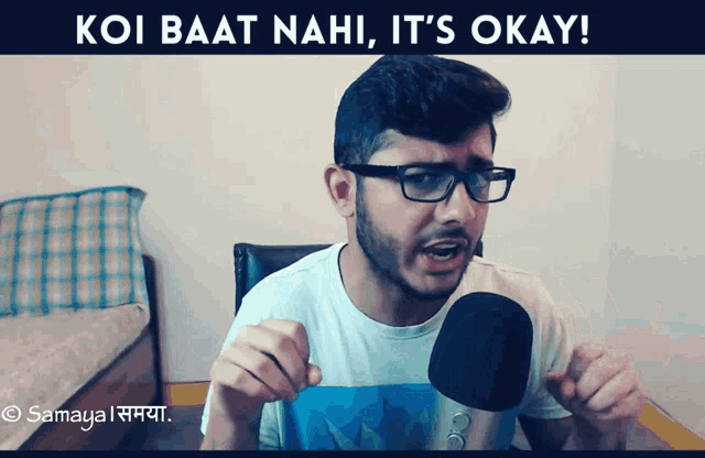 a man with glasses is talking into a microphone with the words koi baat nahi it 's okay written above him