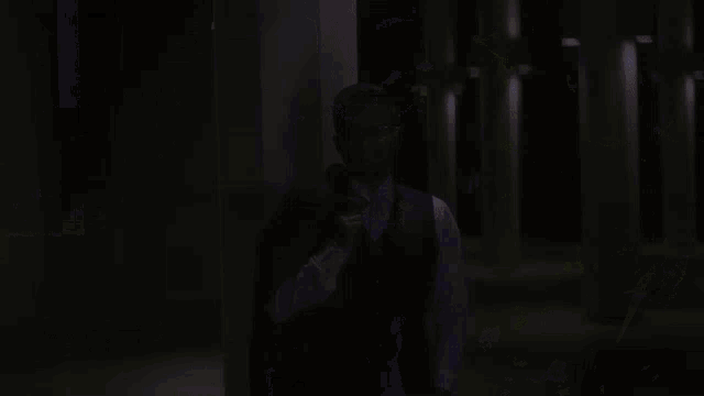 a man wearing glasses and a vest stands in a dark alleyway