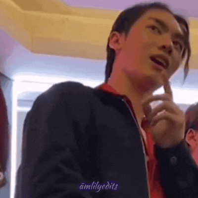 a man in a black jacket and red shirt is standing in a room with his hand on his chin .