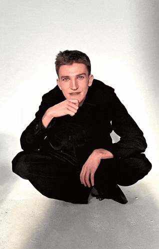 a man in a black jacket is squatting down with his hand on his chin