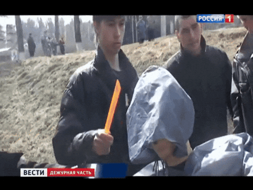 a man in a hooded jacket holds an orange object in front of a screen that says " вести " on it