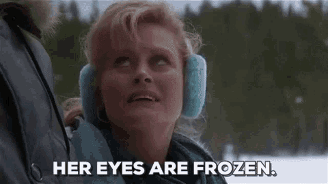 a woman wearing ear muffs and a jacket is standing in the snow and says `` her eyes are frozen '' .
