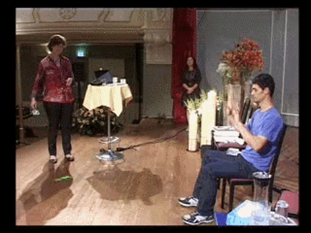 a man in a blue shirt sits in a chair while a woman in a red shirt stands behind him