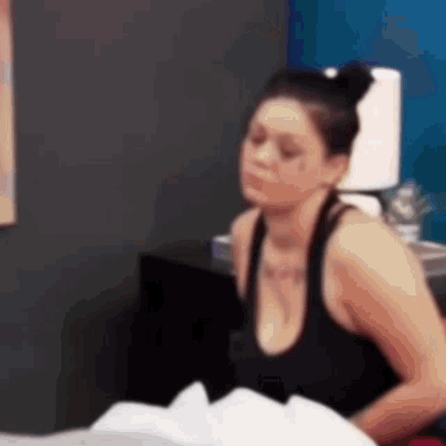 a woman in a black tank top is sitting on a bed looking at something .