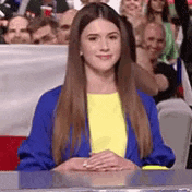 a woman wearing a blue jacket and a yellow shirt is sitting at a table in front of a crowd .