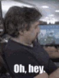 a man in a blue shirt is sitting in front of a computer screen and saying `` oh , hey . ''
