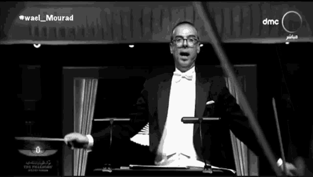 a man in a tuxedo and bow tie is holding a baton in front of a sign that says dmc