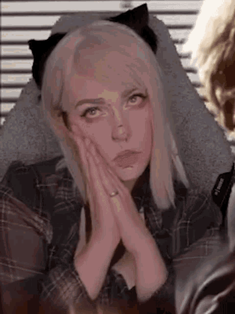 a woman with blonde hair and cat ears is sitting in a chair with her hands folded on her face .