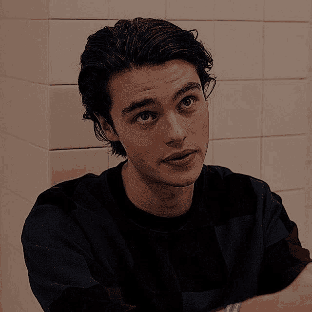 a young man in a black shirt is sitting in front of a white tile wall .