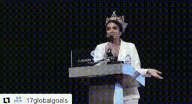 a woman with a crown on her head is standing at a podium speaking into a microphone .