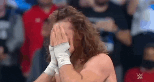 a wrestler with curly hair is covering his face with his hands .